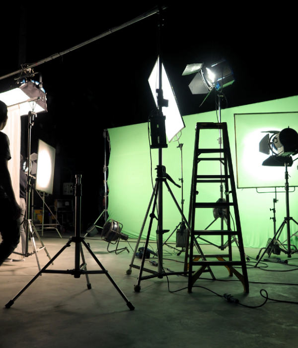 A shadowy atmosphere where two men are walking around with loads of equipment such as ladders, cameras, and a green screen.
