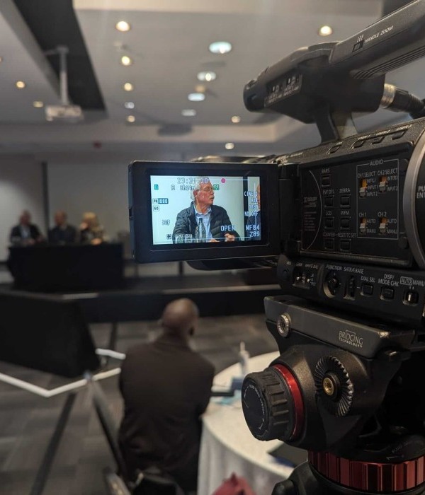 A keynote speaker is visible on a video camera's digital viewfinder and preview screen. Other event attendees and panelists can be seen in the background.